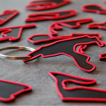 Close-up of multiple black and red 3D-printed Formula 1 track keychains on a dark textured surface with one keychain and metal ring in focus